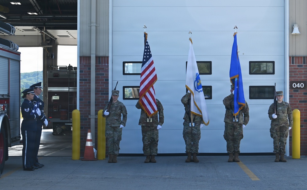 A military color guard ceremony is taking place in front of what appears to be a fire station, with personnel in both dress blues and combat uniforms holding flags. Three flags are displayed: the United States flag, what appears to be the Massachusetts state flag, and a blue military organizational flag. The setting includes a fire station bay with a visible fire truck and yellow bollards.
