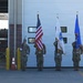 A military color guard ceremony is taking place in front of what appears to be a fire station, with personnel in both dress blues and combat uniforms holding flags. Three flags are displayed: the United States flag, what appears to be the Massachusetts state flag, and a blue military organizational flag. The setting includes a fire station bay with a visible fire truck and yellow bollards.