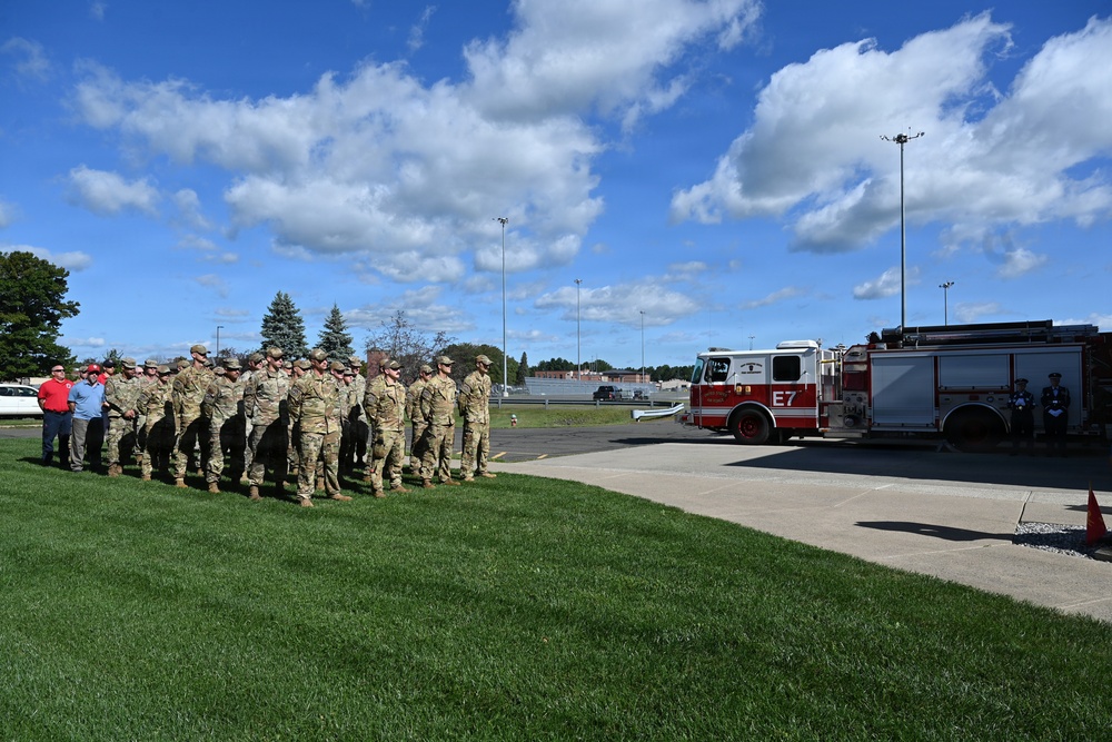 A formation of military personnel in camouflage uniforms stands on a grass area alongside a paved driveway at what appears to be a military installation. A red fire engine labeled 'E7' is parked nearby, with a small group of individuals in darker uniforms standing at attention beside it. The scene suggests a formal ceremony or official event taking place outdoors under partly cloudy skies.
