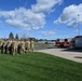 A formation of military personnel in camouflage uniforms stands on a grass area alongside a paved driveway at what appears to be a military installation. A red fire engine labeled 'E7' is parked nearby, with a small group of individuals in darker uniforms standing at attention beside it. The scene suggests a formal ceremony or official event taking place outdoors under partly cloudy skies.