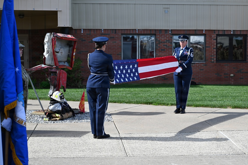 Two U.S. Air Force honor guard members in dress blue uniforms are performing a flag folding ceremony outdoors in front of a brick building. A firefighter in full gear kneels near a twisted metal sculpture or artifact, possibly a 9/11 memorial remnant. A color guard member holding a blue Air Force flag is partially visible on the left side of the frame.