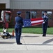 Two U.S. Air Force honor guard members in dress blue uniforms are performing a flag folding ceremony outdoors in front of a brick building. A firefighter in full gear kneels near a twisted metal sculpture or artifact, possibly a 9/11 memorial remnant. A color guard member holding a blue Air Force flag is partially visible on the left side of the frame.