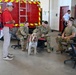 A group of military personnel in camouflage uniforms are gathered inside a fire station bay, interacting with a shaggy dog on a red leash. A civilian in a red shirt and backpack stands nearby, appearing to be the dog's handler. In the background, a red fire/rescue vehicle with yellow chevron markings is visible, along with a person in a white uniform shirt near a doorway.