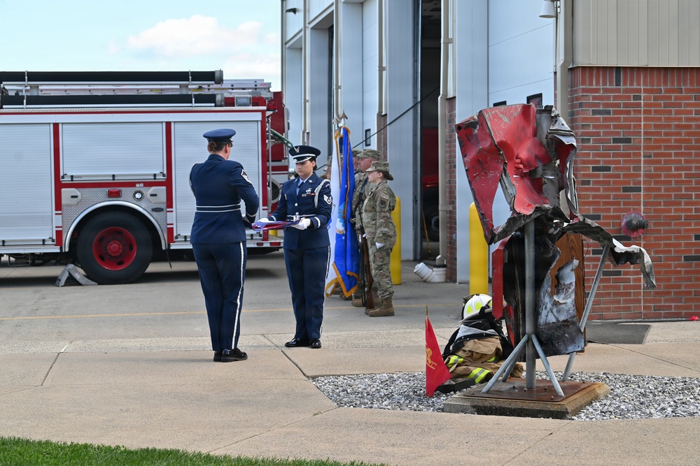 A military ceremony is taking place outside a fire station, with Air Force personnel in dress blues conducting what appears to be a flag-folding or presentation ceremony. A twisted metal sculpture, likely made from debris (possibly 9/11 memorial steel), is displayed in the foreground alongside firefighter gear and a small red flag. Several military personnel in both dress uniforms and combat fatigues stand in formation near the fire station bay.