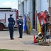A military ceremony is taking place outside a fire station, with Air Force personnel in dress blues conducting what appears to be a flag-folding or presentation ceremony. A twisted metal sculpture, likely made from debris (possibly 9/11 memorial steel), is displayed in the foreground alongside firefighter gear and a small red flag. Several military personnel in both dress uniforms and combat fatigues stand in formation near the fire station bay.