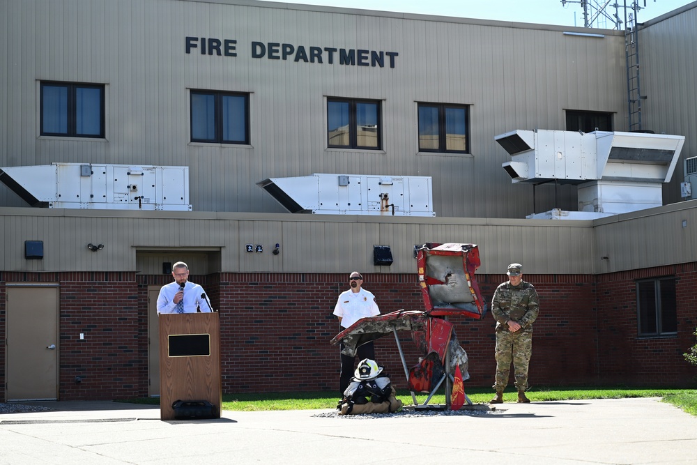An outdoor ceremony or press event is taking place in front of a Fire Department building. A civilian speaker addresses from a wooden podium while a fire department official and a military soldier in camouflage uniform stand nearby. A damaged or burned piece of fire equipment/apparatus is displayed as part of the event.