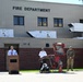 An outdoor ceremony or press event is taking place in front of a Fire Department building. A civilian speaker addresses from a wooden podium while a fire department official and a military soldier in camouflage uniform stand nearby. A damaged or burned piece of fire equipment/apparatus is displayed as part of the event.