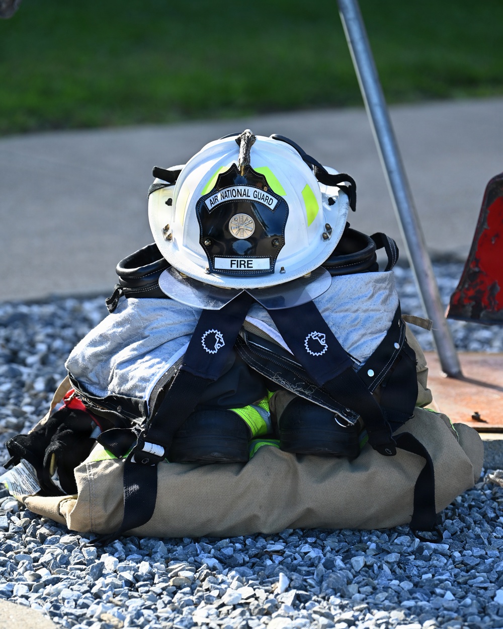 A set of Air National Guard firefighter gear is staged on a gravel surface, ready for rapid donning. The equipment includes a white fire helmet labeled 'AIR NATIONAL GUARD' and 'FIRE,' turnout gear, SCBA harness straps, gloves, and boots, all neatly bundled together. The arrangement suggests pre-positioned gear for a training exercise or emergency response.