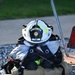 A set of Air National Guard firefighter gear is staged on a gravel surface, ready for rapid donning. The equipment includes a white fire helmet labeled 'AIR NATIONAL GUARD' and 'FIRE,' turnout gear, SCBA harness straps, gloves, and boots, all neatly bundled together. The arrangement suggests pre-positioned gear for a training exercise or emergency response.