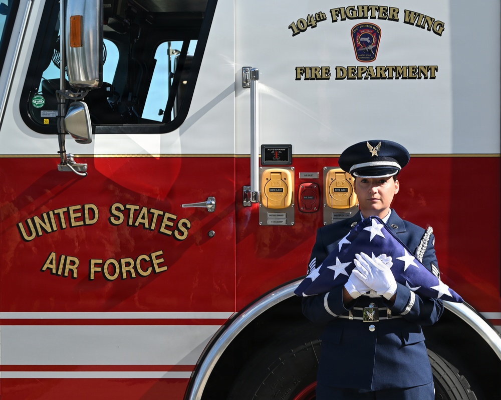 A United States Air Force service member in dress uniform stands at attention in front of a red fire truck belonging to the 104th Fighter Wing Fire Department. The airman is holding a folded American flag with white-gloved hands, suggesting a ceremonial or memorial occasion. The image captures a formal military honor guard moment against the backdrop of a USAF fire apparatus.