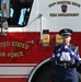 A United States Air Force service member in dress uniform stands at attention in front of a red fire truck belonging to the 104th Fighter Wing Fire Department. The airman is holding a folded American flag with white-gloved hands, suggesting a ceremonial or memorial occasion. The image captures a formal military honor guard moment against the backdrop of a USAF fire apparatus.
