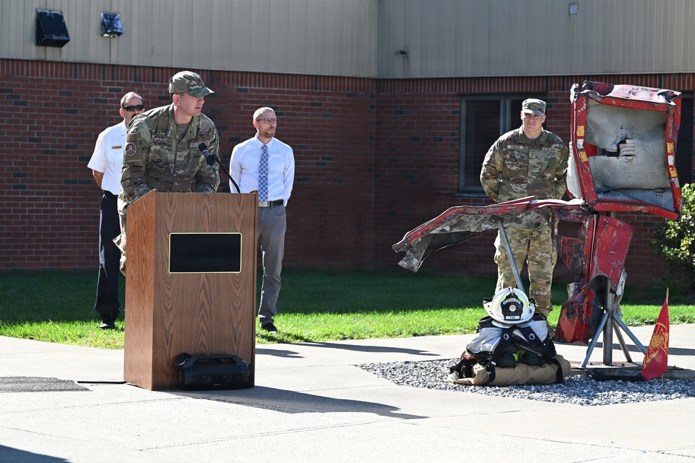 A military ceremony or dedication event is taking place outdoors at what appears to be a military installation. A uniformed service member is speaking at a wooden podium while others stand nearby, with a prominent display featuring damaged/burned red metal structure, firefighting gear including a helmet, and a small flag. The scene suggests a memorial or recognition ceremony, possibly related to firefighting or emergency services.