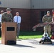 A military ceremony or dedication event is taking place outdoors at what appears to be a military installation. A uniformed service member is speaking at a wooden podium while others stand nearby, with a prominent display featuring damaged/burned red metal structure, firefighting gear including a helmet, and a small flag. The scene suggests a memorial or recognition ceremony, possibly related to firefighting or emergency services.