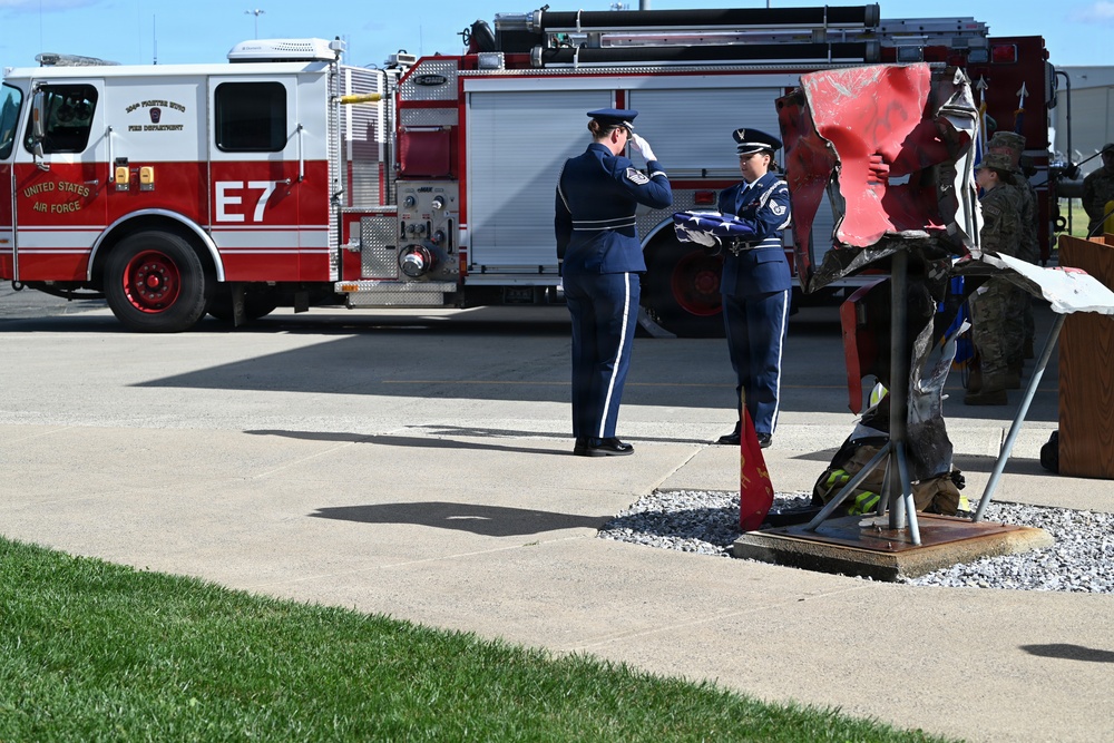 A military ceremony is taking place at a United States Air Force base, featuring two uniformed Air Force personnel performing a flag folding ceremony. A red memorial structure, likely twisted steel remnants, is displayed on a pedestal nearby, suggesting this is a 9/11 memorial or similar commemorative event. A United States Air Force fire truck labeled 'E7' is visible in the background.