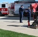 A military ceremony is taking place at a United States Air Force base, featuring two uniformed Air Force personnel performing a flag folding ceremony. A red memorial structure, likely twisted steel remnants, is displayed on a pedestal nearby, suggesting this is a 9/11 memorial or similar commemorative event. A United States Air Force fire truck labeled 'E7' is visible in the background.