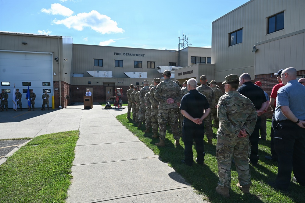 A formal outdoor gathering is taking place in front of a military Fire Department and Civil Engineers building. A large group of personnel in military camouflage uniforms, along with some civilians in plain clothes, are assembled facing a speaker at a podium. The event appears to be an official ceremony or briefing conducted on the building's exterior grounds.