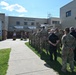 A formal outdoor gathering is taking place in front of a military Fire Department and Civil Engineers building. A large group of personnel in military camouflage uniforms, along with some civilians in plain clothes, are assembled facing a speaker at a podium. The event appears to be an official ceremony or briefing conducted on the building's exterior grounds.
