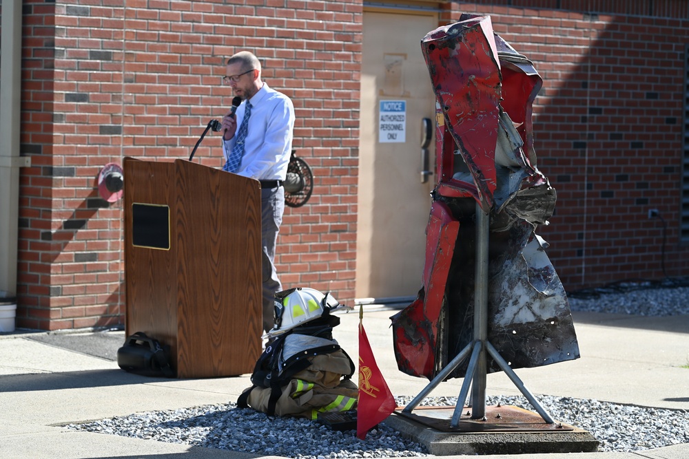 A man in a dress shirt and tie speaks at a wooden podium outdoors, likely at a memorial or dedication ceremony. In the foreground, a sculpture made from crushed/mangled red vehicle parts is mounted on a metal stand, accompanied by firefighter gear and a small red flag. The setting appears to be outside a brick building, possibly a fire station.