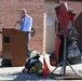 A man in a dress shirt and tie speaks at a wooden podium outdoors, likely at a memorial or dedication ceremony. In the foreground, a sculpture made from crushed/mangled red vehicle parts is mounted on a metal stand, accompanied by firefighter gear and a small red flag. The setting appears to be outside a brick building, possibly a fire station.