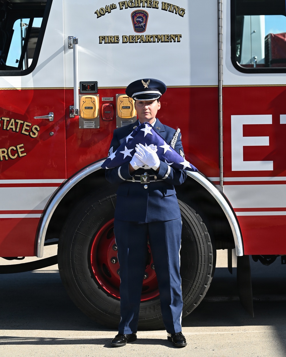 A U.S. Air Force service member in dress blues stands at attention holding a ceremonially folded American flag with white-gloved hands. They are positioned in front of a red fire truck belonging to the 104th Fighter Wing Fire Department. The scene suggests a formal military ceremony, likely a memorial or retirement event.