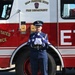 A U.S. Air Force service member in dress blues stands at attention holding a ceremonially folded American flag with white-gloved hands. They are positioned in front of a red fire truck belonging to the 104th Fighter Wing Fire Department. The scene suggests a formal military ceremony, likely a memorial or retirement event.