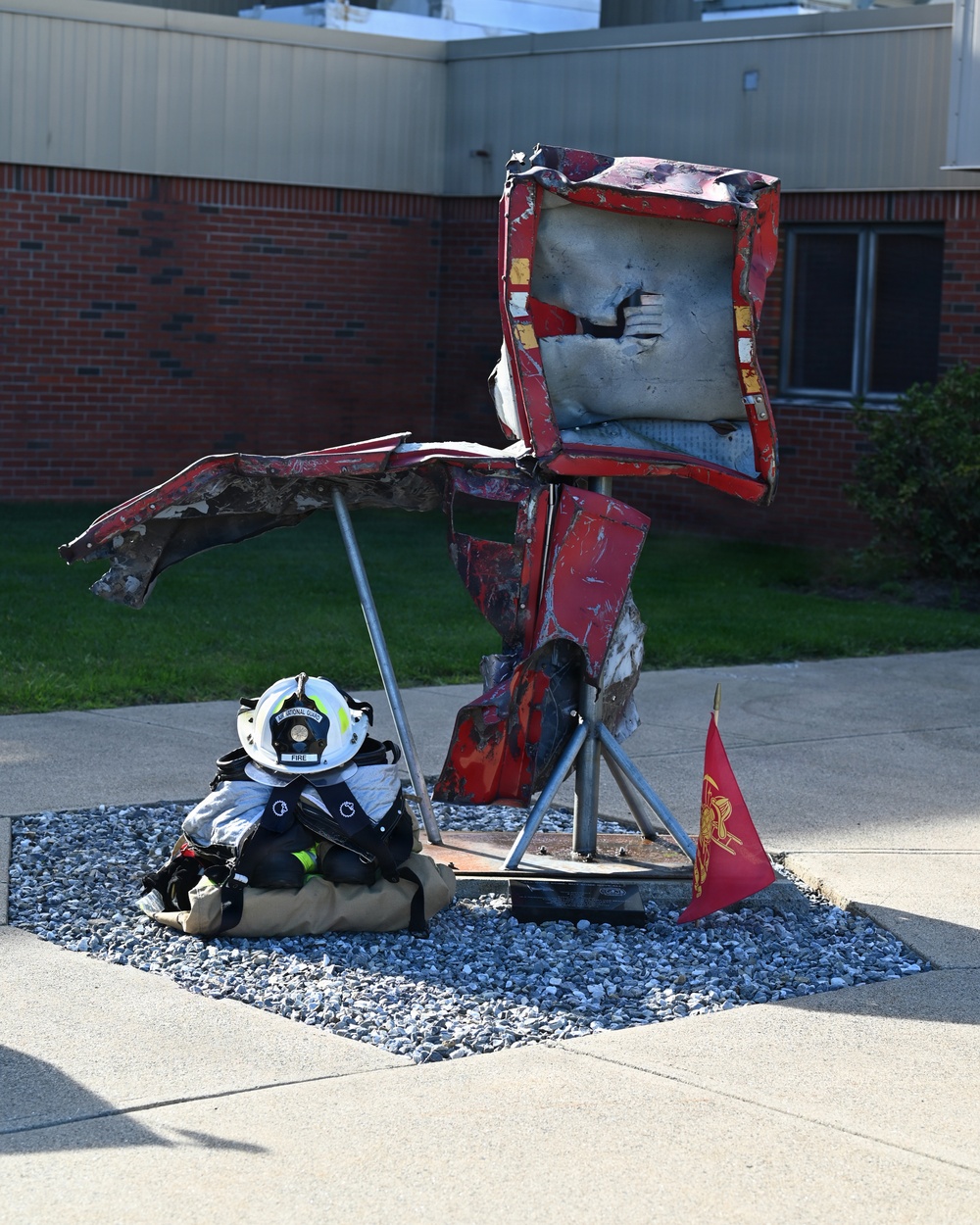 A firefighter in full gear, including a white helmet marked 'FIRE', is seated or crouching on a gravel bed beside a memorial sculpture made from twisted, burned red metal debris. The sculpture appears to be constructed from wreckage — possibly from a fire truck or emergency vehicle — mounted on a metal stand. A small red flag bearing a firefighter emblem is placed at the base of the sculpture, suggesting this is a memorial or tribute display outside a brick building.