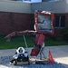 A firefighter in full gear, including a white helmet marked 'FIRE', is seated or crouching on a gravel bed beside a memorial sculpture made from twisted, burned red metal debris. The sculpture appears to be constructed from wreckage — possibly from a fire truck or emergency vehicle — mounted on a metal stand. A small red flag bearing a firefighter emblem is placed at the base of the sculpture, suggesting this is a memorial or tribute display outside a brick building.