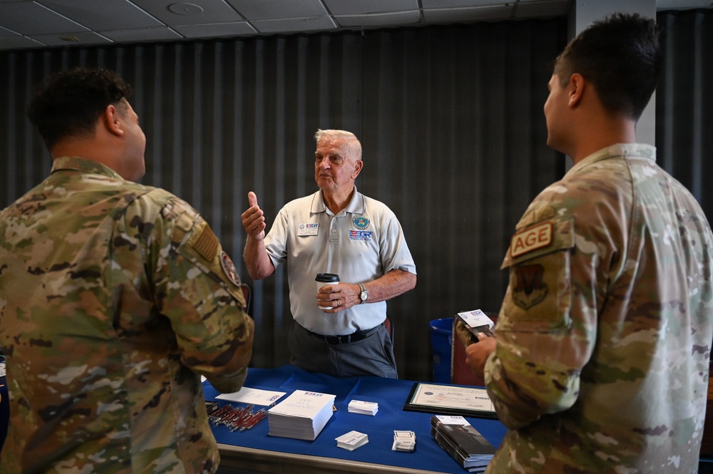An elderly civilian man wearing an ESGR (Employer Support of the Guard and Reserve) polo shirt is speaking with two U.S. Air Force military personnel in OCP (Operational Camouflage Pattern) uniforms at an informational table. The table is covered with a blue cloth and displays brochures, business cards, and framed certificates. The setting appears to be an indoor event or outreach booth.