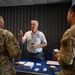 An elderly civilian man wearing an ESGR (Employer Support of the Guard and Reserve) polo shirt is speaking with two U.S. Air Force military personnel in OCP (Operational Camouflage Pattern) uniforms at an informational table. The table is covered with a blue cloth and displays brochures, business cards, and framed certificates. The setting appears to be an indoor event or outreach booth.
