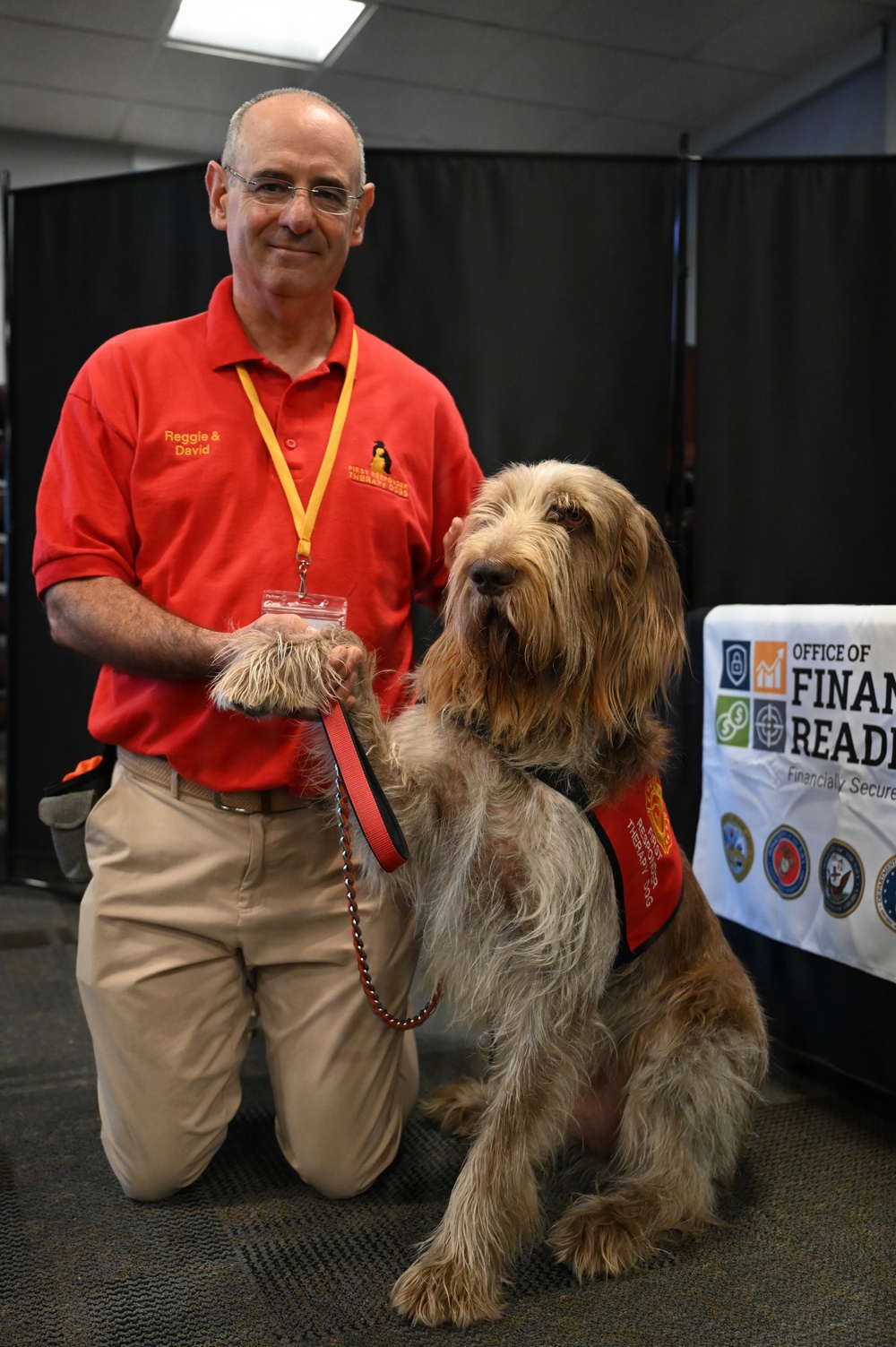 A man named David, wearing a red polo shirt with 'Reggie &amp; David' embroidered on it, kneels beside a large shaggy therapy dog named Reggie at what appears to be an indoor event or expo. The dog is wearing a red therapy dog vest and is holding up its paw toward the man's hand. A partially visible banner for the 'Office of Financial Readiness' is seen in the background.