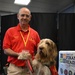 A man named David, wearing a red polo shirt with 'Reggie &amp; David' embroidered on it, kneels beside a large shaggy therapy dog named Reggie at what appears to be an indoor event or expo. The dog is wearing a red therapy dog vest and is holding up its paw toward the man's hand. A partially visible banner for the 'Office of Financial Readiness' is seen in the background.