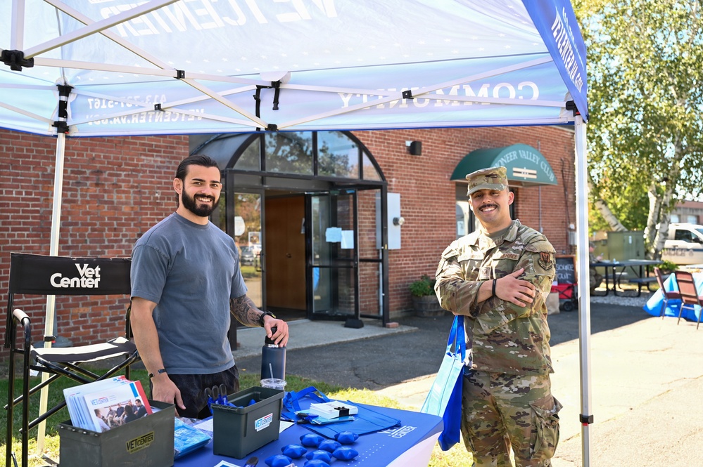 Two men are standing at an outdoor Vet Center community outreach booth, smiling at the camera. One man is in civilian clothing while the other is wearing a U.S. Air Force military uniform in OCP (Operational Camouflage Pattern). The event appears to be taking place in a parking lot in front of a brick building identified as the Pioneer Valley Club.