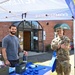 Two men are standing at an outdoor Vet Center community outreach booth, smiling at the camera. One man is in civilian clothing while the other is wearing a U.S. Air Force military uniform in OCP (Operational Camouflage Pattern). The event appears to be taking place in a parking lot in front of a brick building identified as the Pioneer Valley Club.