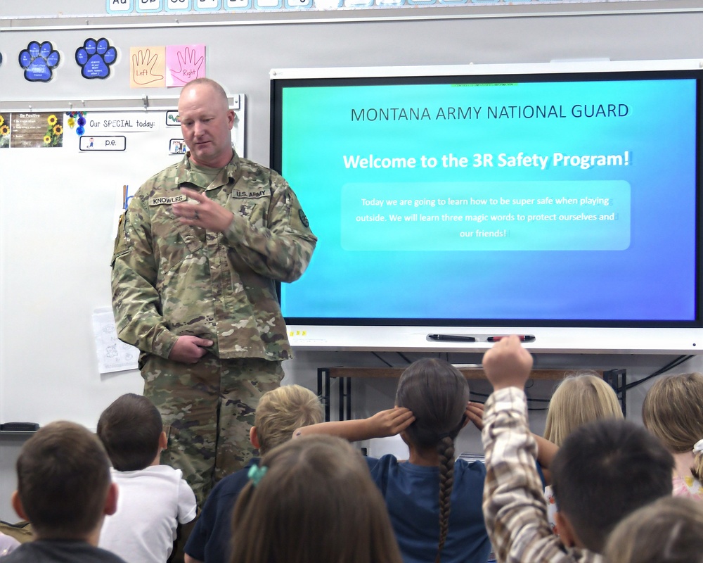 A U.S. Army soldier named Knowles, wearing OCP camouflage uniform, is presenting the Montana Army National Guard's '3R Safety Program' to a classroom of young elementary school children. The presentation is displayed on a large interactive smartboard, welcoming students to learn safety skills for playing outside. Several children are engaged, with at least one raising their hand.