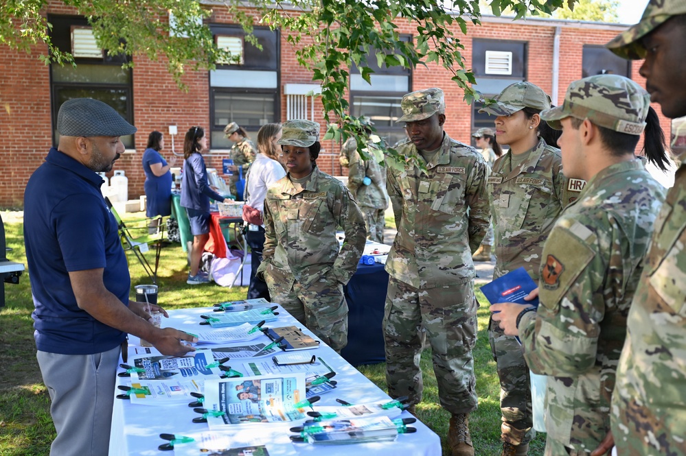 A group of U.S. Air Force personnel in OCP (Operational Camouflage Pattern) uniforms are attending an outdoor information or resource fair near a brick building. A civilian man in a navy polo shirt is presenting materials at a table covered with brochures, lanyards, and informational pamphlets. The event appears to be a wellness, benefits, or community outreach fair held on a military installation.
