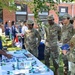 A group of U.S. Air Force personnel in OCP (Operational Camouflage Pattern) uniforms are attending an outdoor information or resource fair near a brick building. A civilian man in a navy polo shirt is presenting materials at a table covered with brochures, lanyards, and informational pamphlets. The event appears to be a wellness, benefits, or community outreach fair held on a military installation.