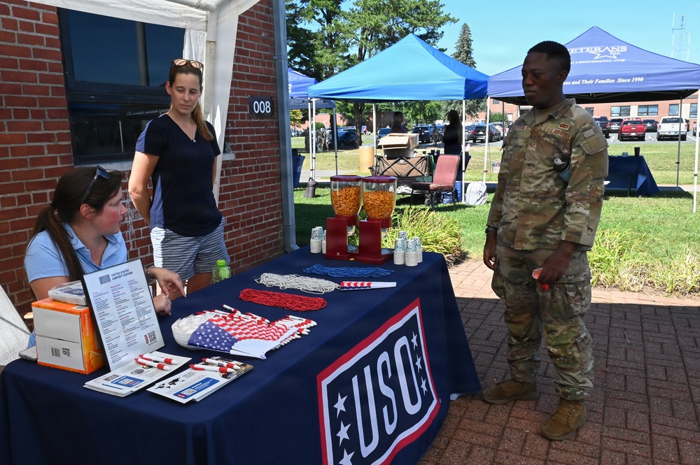 A USO booth is set up outdoors at what appears to be a military installation event. A seated female volunteer and a standing civilian woman interact with a U.S. Army soldier in OCP camouflage uniform. The table displays patriotic items including American flag decorations, red/white/blue bead necklaces, candy dispensers, and informational brochures.