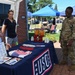 A USO booth is set up outdoors at what appears to be a military installation event. A seated female volunteer and a standing civilian woman interact with a U.S. Army soldier in OCP camouflage uniform. The table displays patriotic items including American flag decorations, red/white/blue bead necklaces, candy dispensers, and informational brochures.