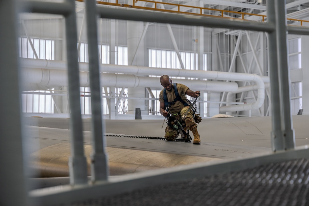 A military personnel member wearing camouflage uniform and a safety harness is working on top of what appears to be a large aircraft inside a spacious hangar facility. The individual is crouching and handling a chain or cable while secured with fall-protection equipment. The photo is taken through a metal grating or railing structure in the foreground, giving a framed perspective.