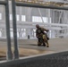 A military personnel member wearing camouflage uniform and a safety harness is working on top of what appears to be a large aircraft inside a spacious hangar facility. The individual is crouching and handling a chain or cable while secured with fall-protection equipment. The photo is taken through a metal grating or railing structure in the foreground, giving a framed perspective.