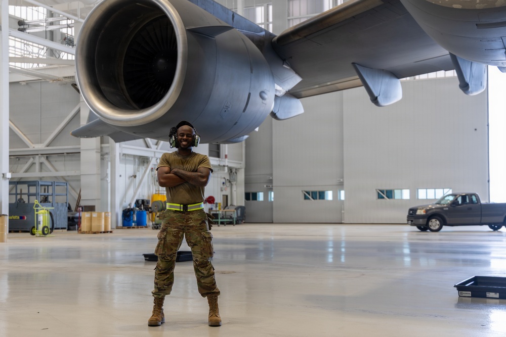A military aircraft maintainer stands confidently with arms crossed beneath a large jet engine inside a military hangar. The individual is wearing OCP (Operational Camouflage Pattern) uniform, hearing protection, and a high-visibility safety vest, smiling broadly at the camera. The setting is a well-lit, spacious aircraft maintenance hangar with support equipment and a pickup truck visible in the background.