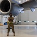 A military aircraft maintainer stands confidently with arms crossed beneath a large jet engine inside a military hangar. The individual is wearing OCP (Operational Camouflage Pattern) uniform, hearing protection, and a high-visibility safety vest, smiling broadly at the camera. The setting is a well-lit, spacious aircraft maintenance hangar with support equipment and a pickup truck visible in the background.