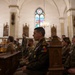 Military personnel from what appears to be both U.S. Army and Polish Army are seated in church pews during a religious ceremony or memorial service. The setting is a Gothic-style Christian church with stained glass windows, vaulted ceilings, and ornate wooden furnishings. The central figure wears U.S. Army dress uniform with visible patches including 'Ranger' and 'Old Ironsides' (1st Armored Division).