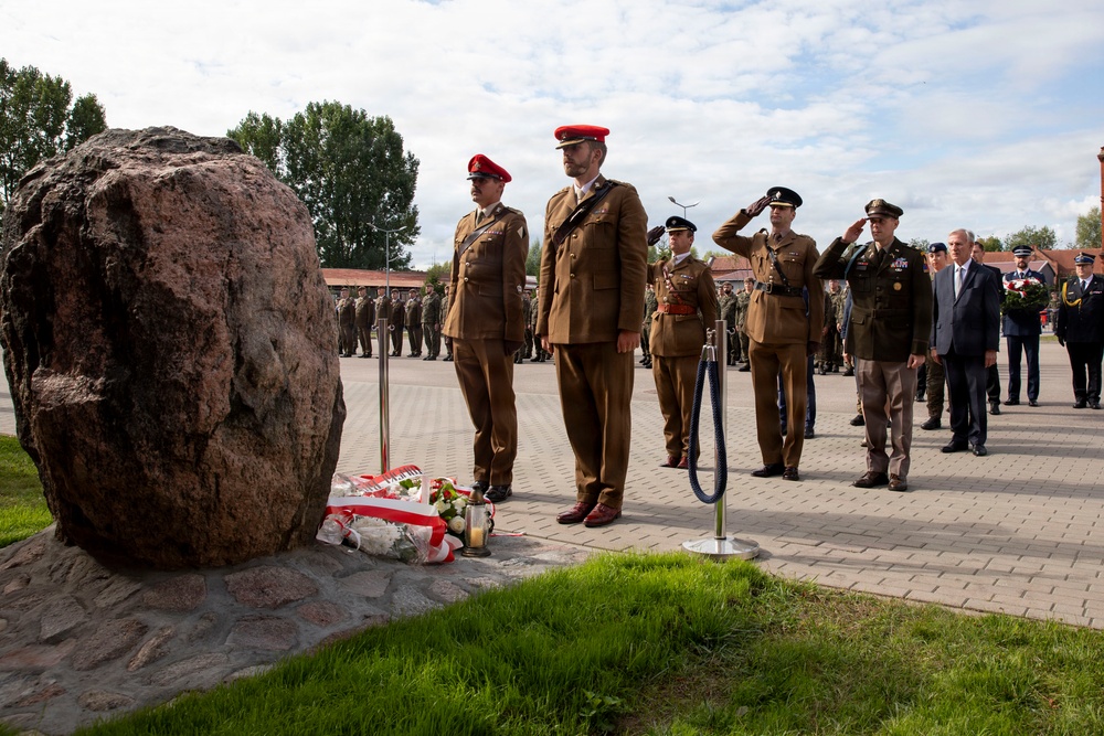 A military ceremony is taking place at an outdoor memorial site featuring a large natural boulder monument with flowers and a candle placed at its base. Several military personnel in brown uniforms with red berets, along with others in different uniforms, are standing at attention or saluting. A civilian in a suit is also present among the group, suggesting an official commemorative event.
