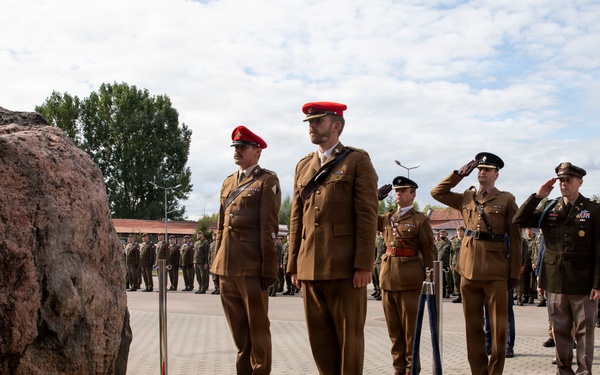 A military ceremony is taking place at an outdoor memorial site featuring a large natural boulder monument with flowers and a candle placed at its base. Several military personnel in brown uniforms with red berets, along with others in different uniforms, are standing at attention or saluting. A civilian in a suit is also present among the group, suggesting an official commemorative event.