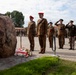 A military ceremony is taking place at an outdoor memorial site featuring a large natural boulder monument with flowers and a candle placed at its base. Several military personnel in brown uniforms with red berets, along with others in different uniforms, are standing at attention or saluting. A civilian in a suit is also present among the group, suggesting an official commemorative event.