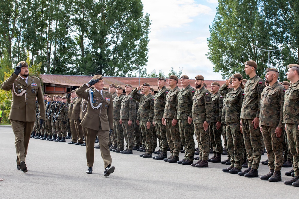 A military parade or inspection ceremony is taking place outdoors on a paved surface. Two senior officers in dress uniforms are walking and saluting while reviewing a formation of soldiers standing at attention in camouflage uniforms. The soldiers display Polish flag patches on their shoulders, indicating this is a Polish Armed Forces event.