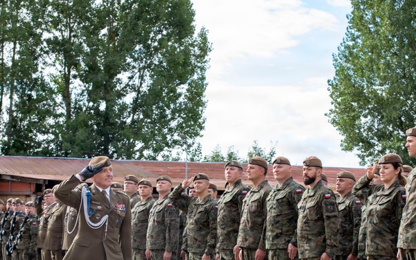 A military parade or inspection ceremony is taking place outdoors on a paved surface. Two senior officers in dress uniforms are walking and saluting while reviewing a formation of soldiers standing at attention in camouflage uniforms. The soldiers display Polish flag patches on their shoulders, indicating this is a Polish Armed Forces event.