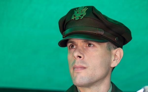 A U.S. Army officer in Army Service Uniform (ASU) is photographed against a green backdrop. The officer, identified by nameplate as 'MARQUIS,' appears to be engaged in conversation or a briefing, with hands clasped together. A second individual in camouflage uniform is partially visible at the right edge of the frame.