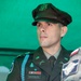 A U.S. Army officer in Army Service Uniform (ASU) is photographed against a green backdrop. The officer, identified by nameplate as 'MARQUIS,' appears to be engaged in conversation or a briefing, with hands clasped together. A second individual in camouflage uniform is partially visible at the right edge of the frame.